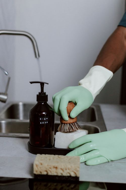 Handen met groene handschoenen wassen af met afwasmiddel en een borstel in de spoelbak